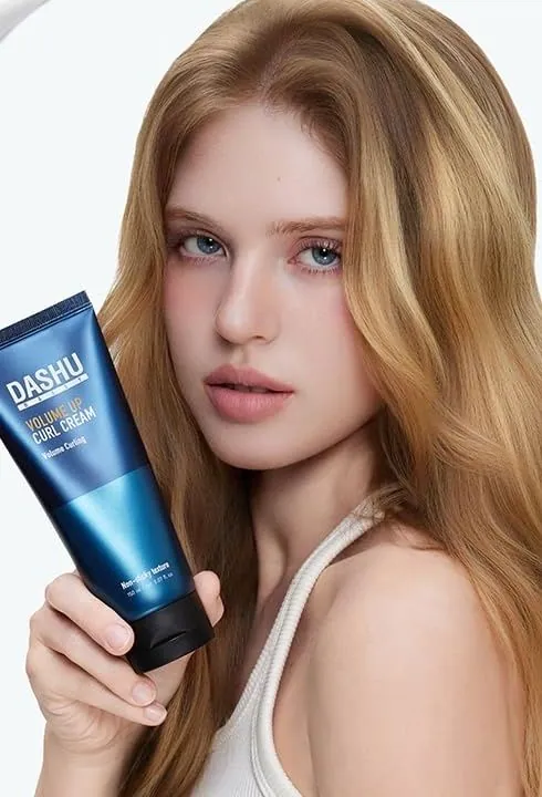 A model with long wavy hair holding a blue tube of DASHU Daily Volume Up Curl Cream
