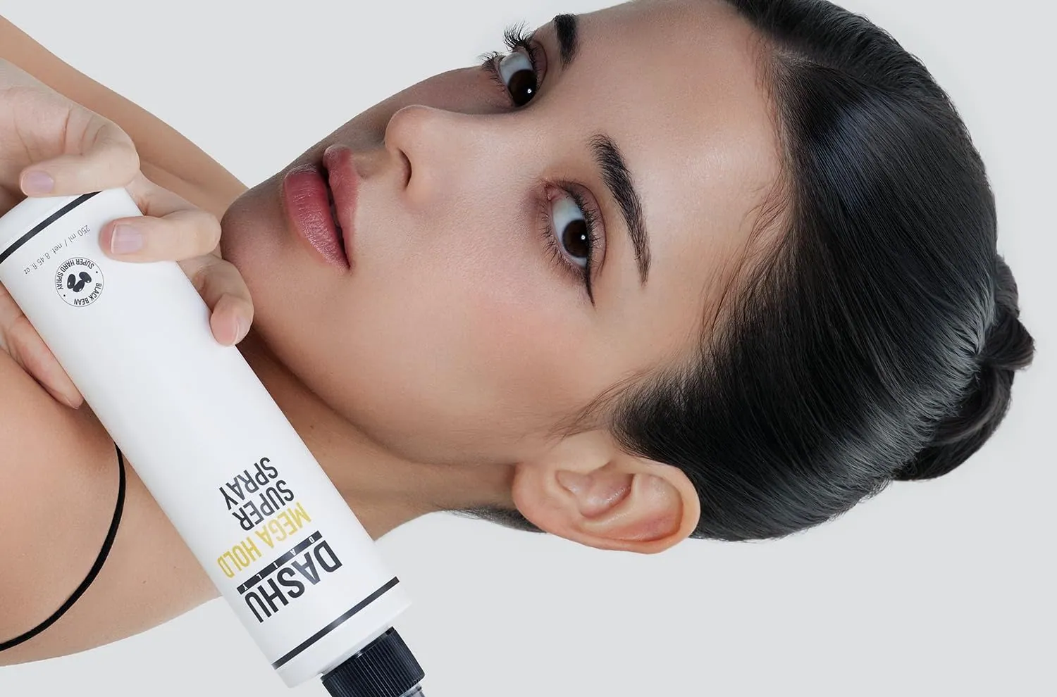 A woman with a slick bun holding a bottle of DASHU Mega Hold Super Hair Spray